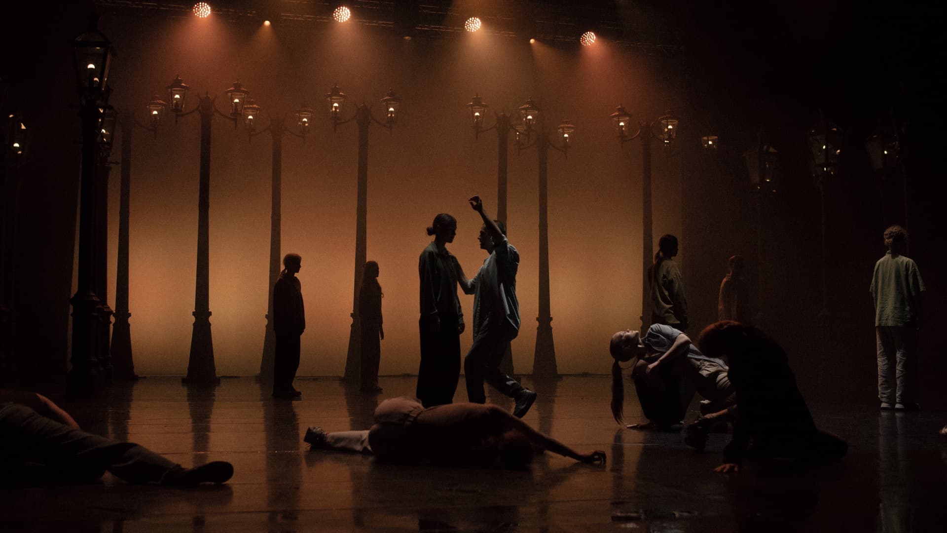Performers forming a layered stage composition beneath warm amber lights and rows of street lamps at Green Theatre.