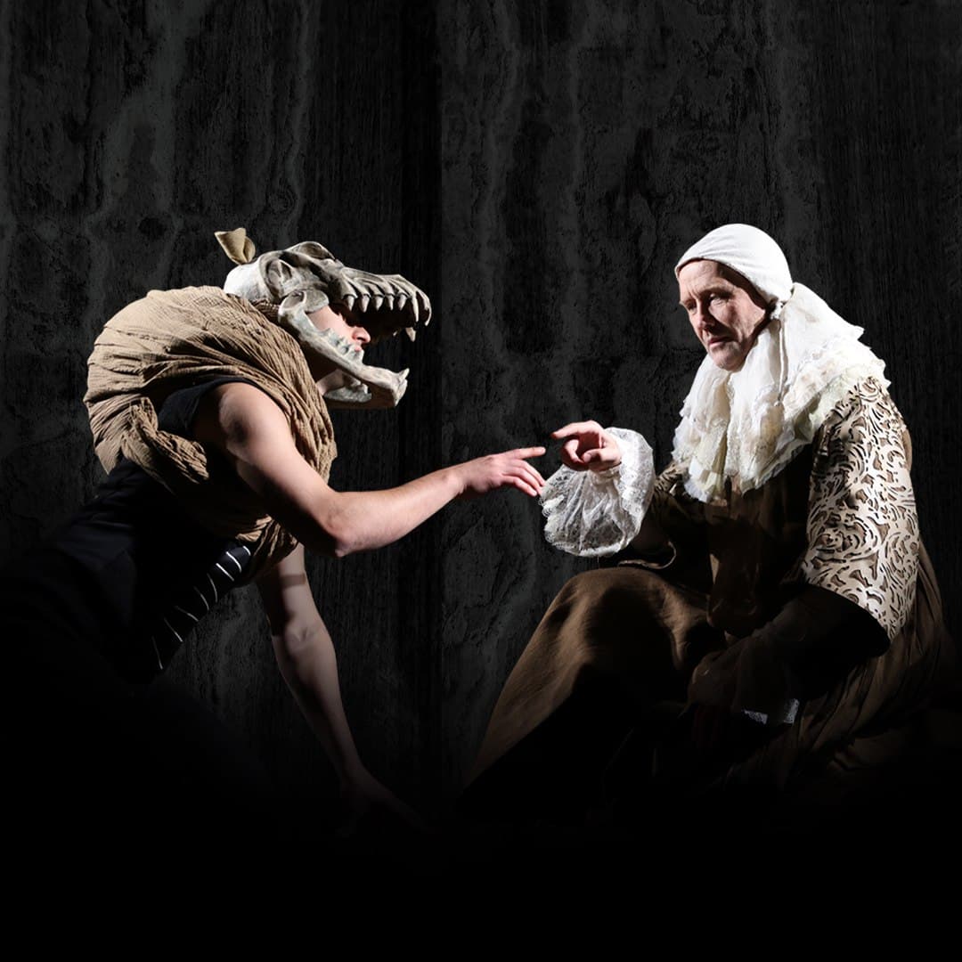 Mephistopheles in a dog mask reaches toward Faust under hard stage light.